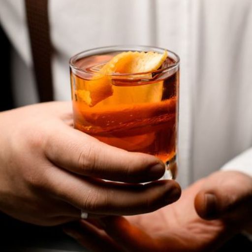 Bartender in white shirt holding a clear cocktial with orange garnish.