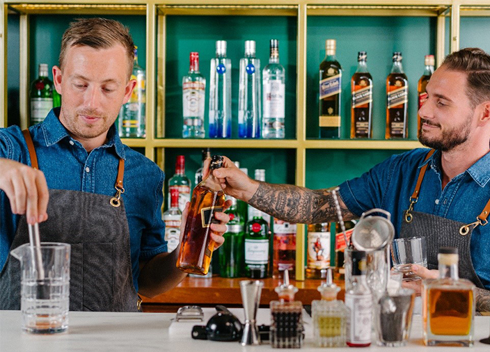 Two male DBA bartenders behind the bar working together. Bartender on the right offer a bottle of Johnnie Walker Black Label to his colleague who is looking at stirring a cocktail.