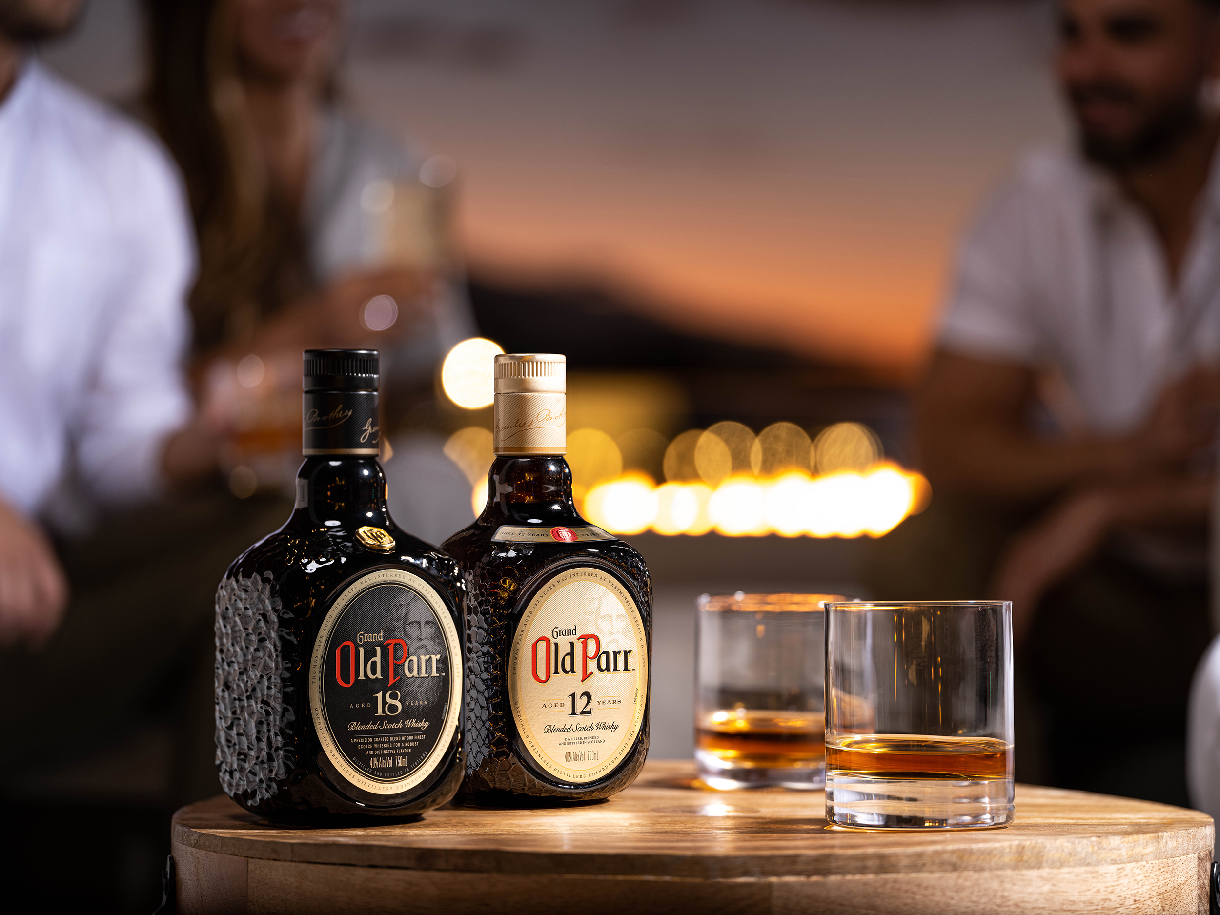 Garrafas de whisky Grand Old Parr 18 e 12 anos sobre mesa de madeira redonda, ao lado de dois copos baixos com bebida. Fundo desfocado mostra pessoas em ambiente social ao entardecer, com iluminação quente e luzes bokeh.