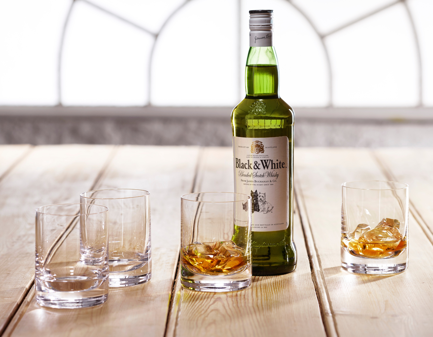 Bottle of Black & White whisky on wooden table surrounded by whisky tumblers