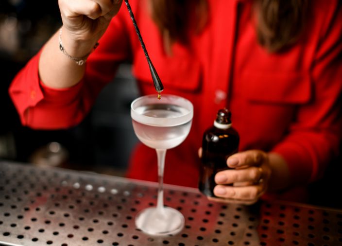 A bartender dropping a brown liquid into a chilled glass of clear liquid.