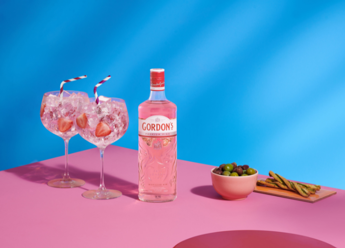 Botella de gin Premium Pink de Gordon's junto a dos vasos de G&T frente a un fondo rosa y azul.