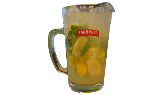 Jarra de vidro com logo vermelho da Smirnoff contendo um coquetel refrescante amarelo-claro. A bebida está cheia de cubos de gelo e pedaços rústicos de limão verde e limão siciliano misturados no interior. Fundo branco.