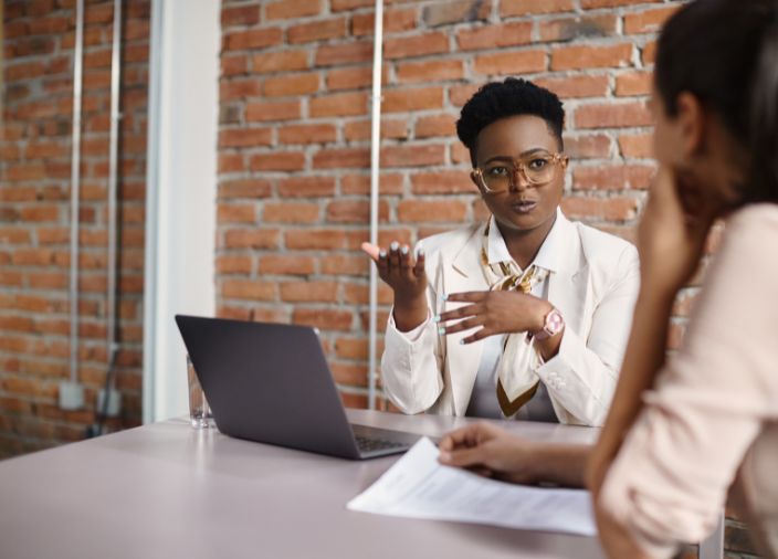 Un mentor habla de tú a tú con un miembro del personal mientras está sentado en un escritorio con un ordenador portátil delante.