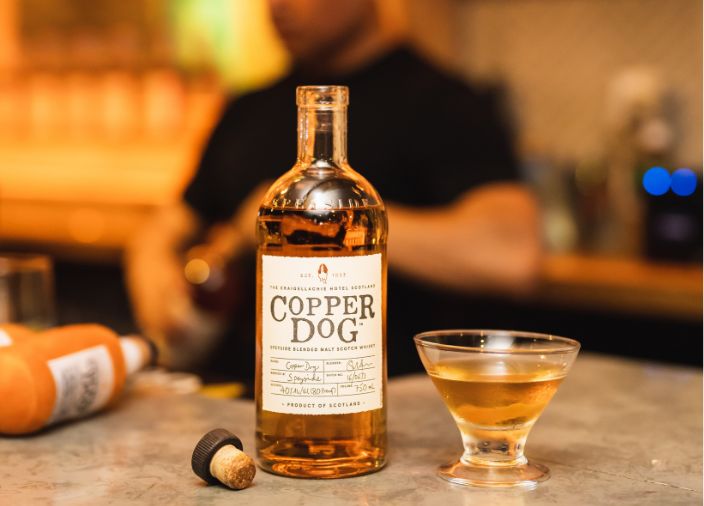 Una botella abierta de Copper Dog con un vaso de whisky puro, el bartender al fondo
