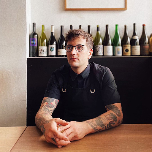Head Chef del Bar Liberty de Melbourne, Zack Leon Furst, sentado en una mesa con las manos juntas, mirando hacia la izquierda de la pantalla. Una hilera de botellas de vino detrás de él.