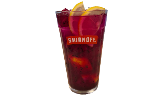Copo alto com logo vermelho da Smirnoff contendo um coquetel rosa escuro vibrante, característico de pitaya, com sementinhas pretas visíveis misturadas no líquido. O topo da bebida é decorado com duas fatias de limão siciliano. Fundo branco.