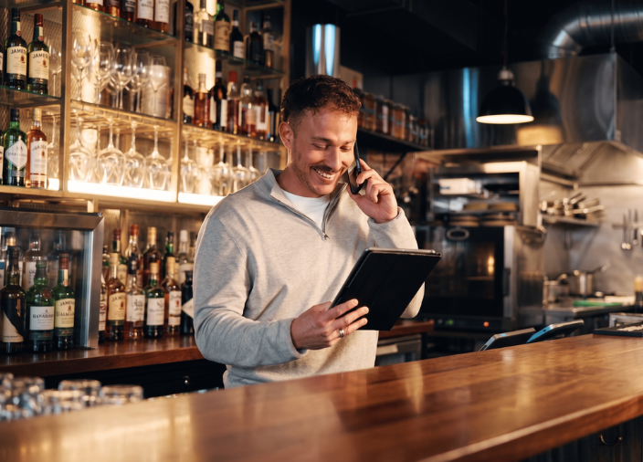 Homem sorridente atrás de um balcão de bar em madeira. Ele fala ao celular enquanto segura e olha para um tablet. Ao fundo, prateleiras iluminadas repletas de garrafas e taças, além de uma cozinha industrial desfocada.
