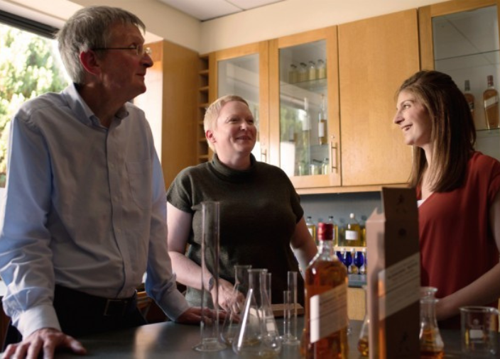 Three people gathered in kitchen around whisky bottle and glasses.    
