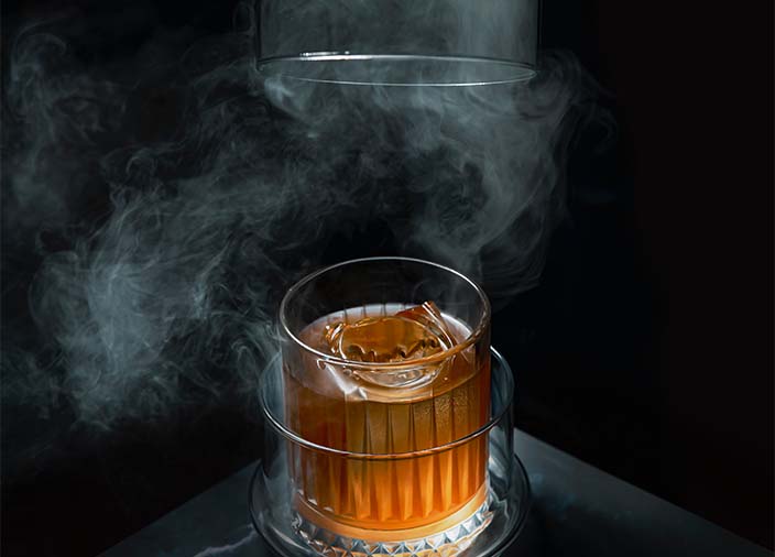 Ein gefülltes Glas mit Flüssigkeit, umgeben von Rauch vor einem schwarzen Hintergrund.