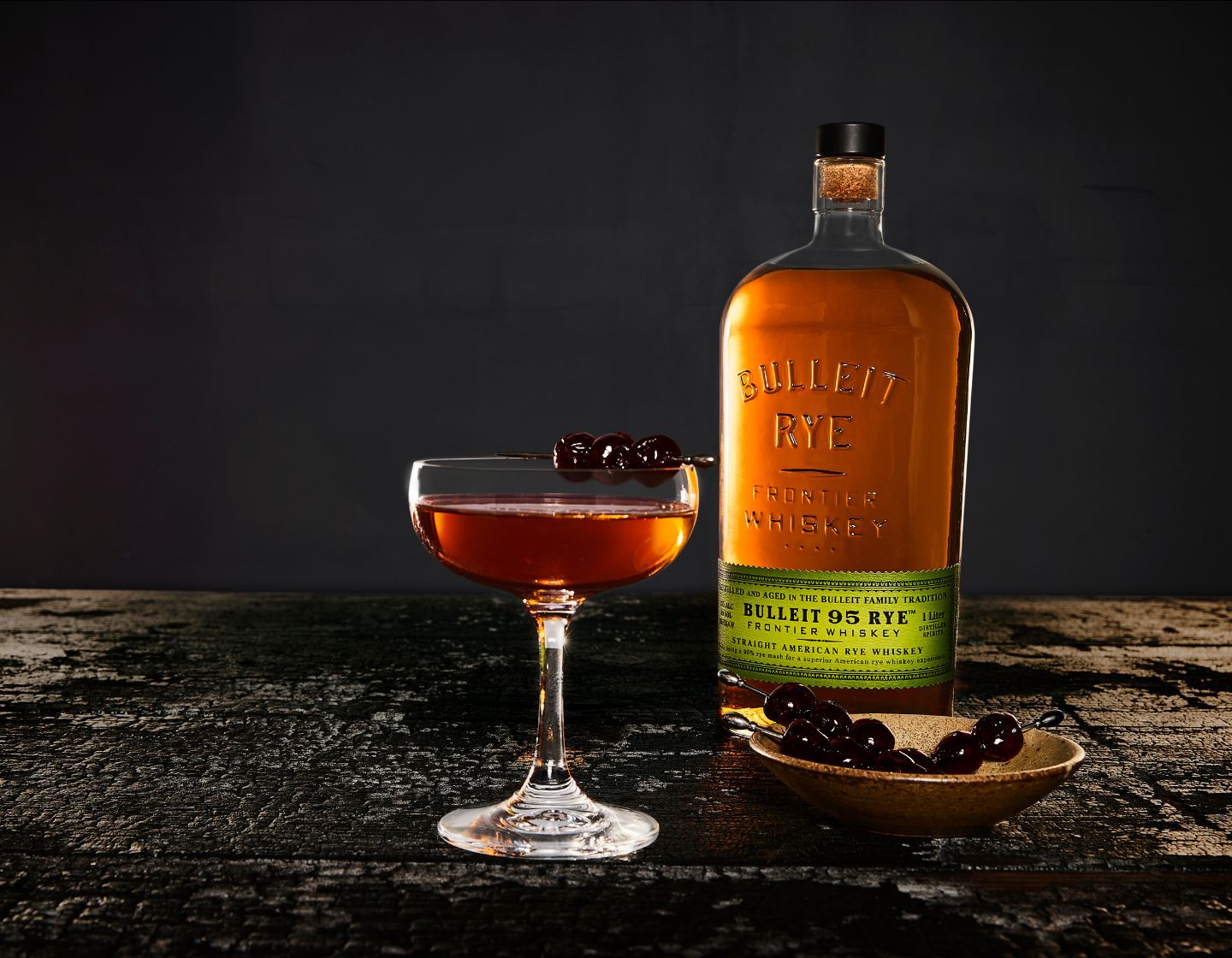 Garrafa de bourbon Bulleit ao lado de um copo de coquetel de whiskey decorado com cerejas