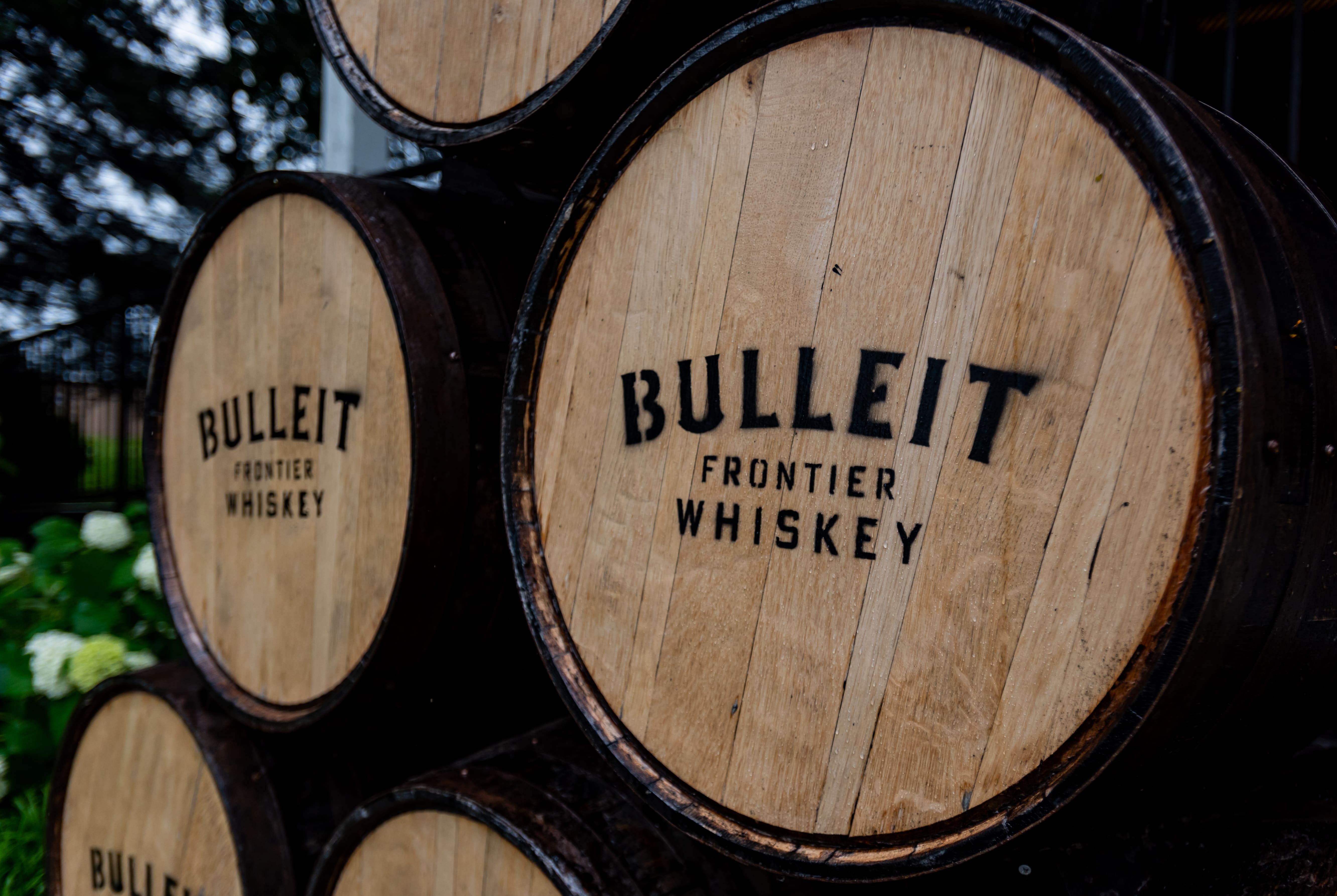 Barris de madeira rústica empilhados. A face do barril em destaque exibe a marca 'BULLEIT FRONTIER WHISKEY' estampada em letras pretas. O fundo levemente desfocado mostra folhagens escuras, sugerindo uma destilaria ao ar livre.