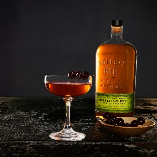 Bulleit_bourbon_bottle_beside_coupe_of_whiskey_cocktail_garnished_with_cherries