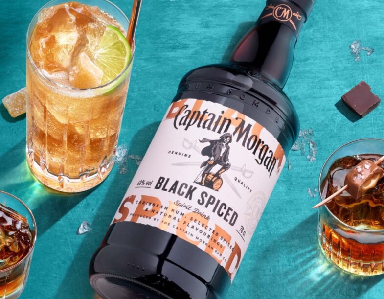 Bottle of Captain Morgan Black Spiced Rum on blue background beside a short glass of dark liquid and a tall glass of gold liquid.