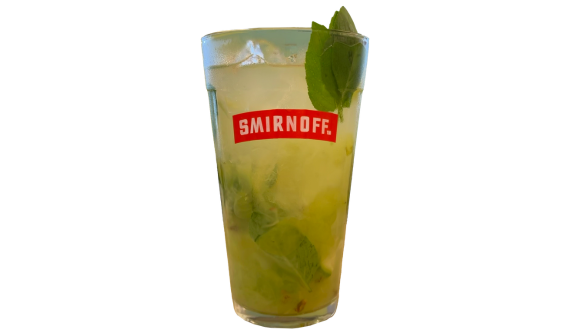 Copo alto com logo vermelho da Smirnoff contendo um coquetel verde-claro e turvo. A bebida refrescante é misturada com gelo e folhas verdes, sendo decorada no topo por um generoso ramo de manjericão fresco. Fundo branco isolado.