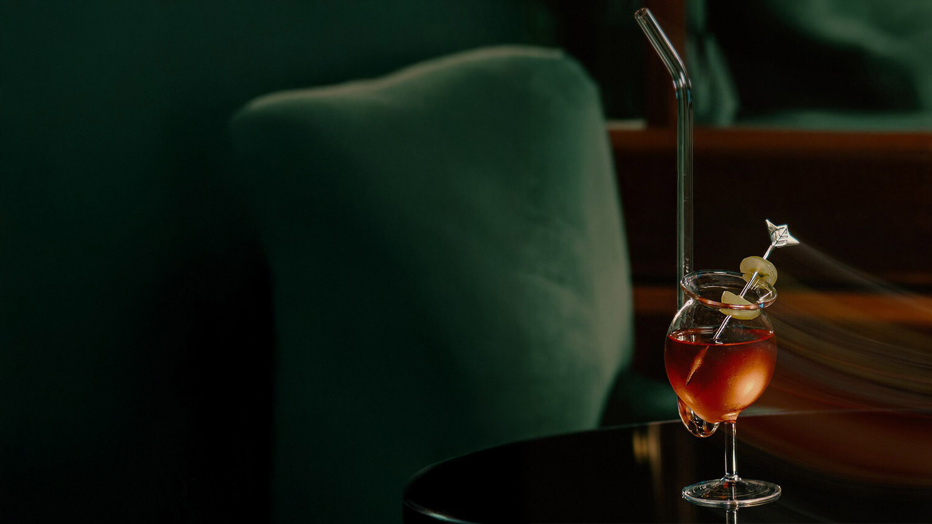 Coquetel avermelhado em taça de vidro com bojo esférico e haste curta, sobre mesa escura reflexiva. A bebida é decorada com um canudo de vidro longo e um palito prateado espetando uma guarnição redonda. Fundo verde-escuro desfocado e elegante.