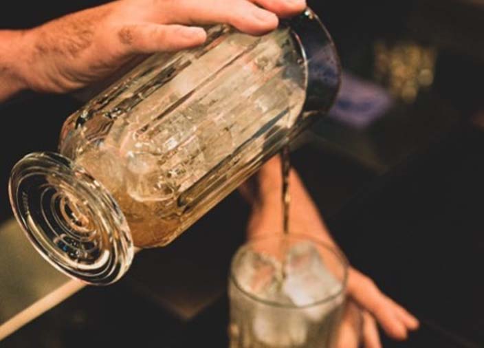 A bartender uses a mixer with a stirring spoon to pour a cocktail mix into a glass of ice.