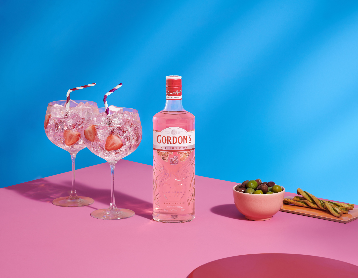 Botella de ginebra Gordon’s Premium Pink junto a dos copas de gin-tonic sobre un fondo rosa y azul