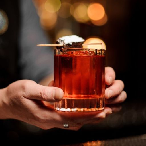 A person holding a red cocktail with a brownie garnish topped with icing sugar.