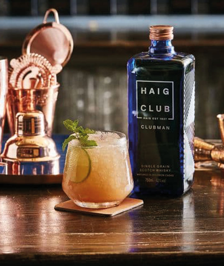 Botella Haig Club al lado un vaso servido y decorado de menta y alrededor utensilios