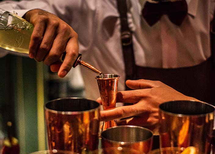 Close up of a person wearing a bow tie and apron pouring liquid into a jigger.