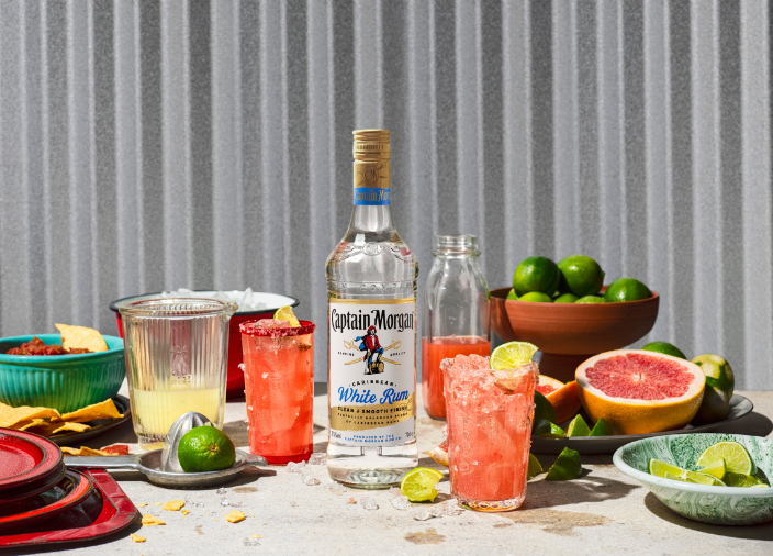 Bottle of Captain Morgan White Rum on table surrounded by assorted food and drink against white background