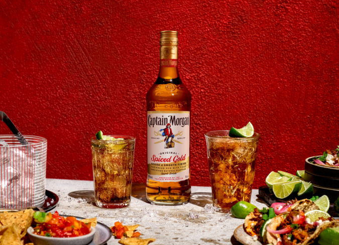 Botella de ron Captain Morgan Spiced Gold sobre una mesa junto a platos de comida frente a un fondo rojo.