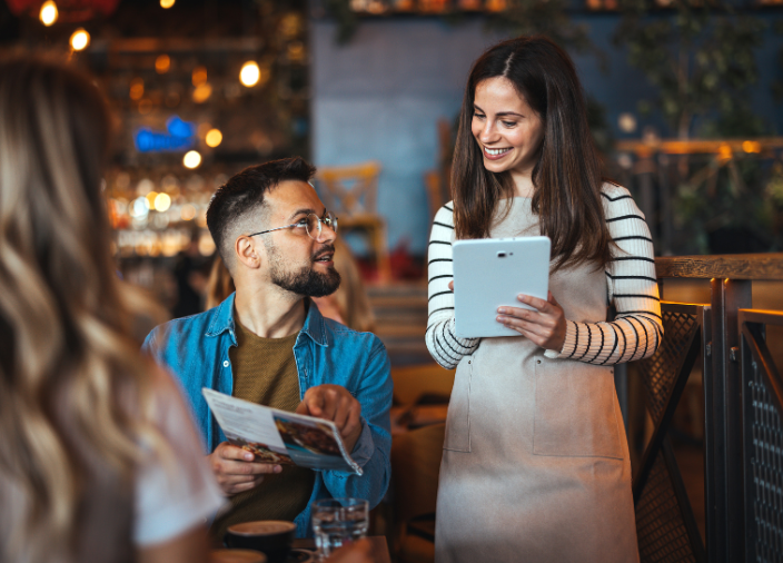 Garçonete sorridente de avental anota o pedido em um tablet. O cliente, um homem de óculos, olha para ela enquanto aponta para o cardápio aberto em suas mãos. Ambiente de restaurante acolhedor com iluminação quente.