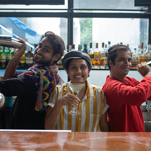 Three bartenders posed together, those on the outside holding cocktail shakers and smiling at the camera. The one in the middle lifting a cocktail glass and smiling at the camera. 