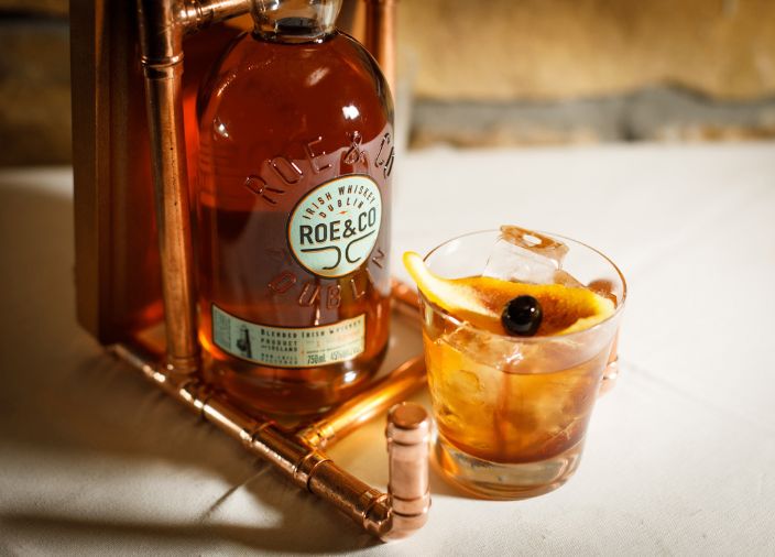 A Roe & Red cocktail with an orange slice garnish next to a bottle of Roe & Co