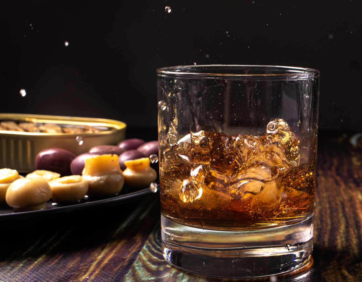   Vaso de whisky con hielo con plato de aceitunas y nueces detrás
