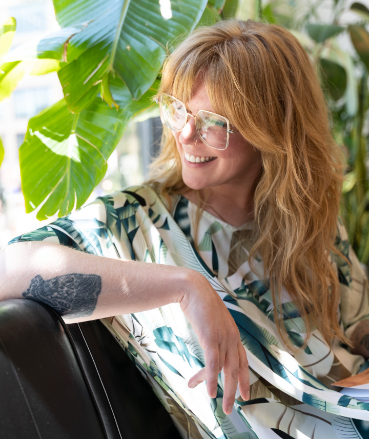 Laura Green sitting outside on a chair and smiling. Wearing top with plant print, glasses and long hair. 