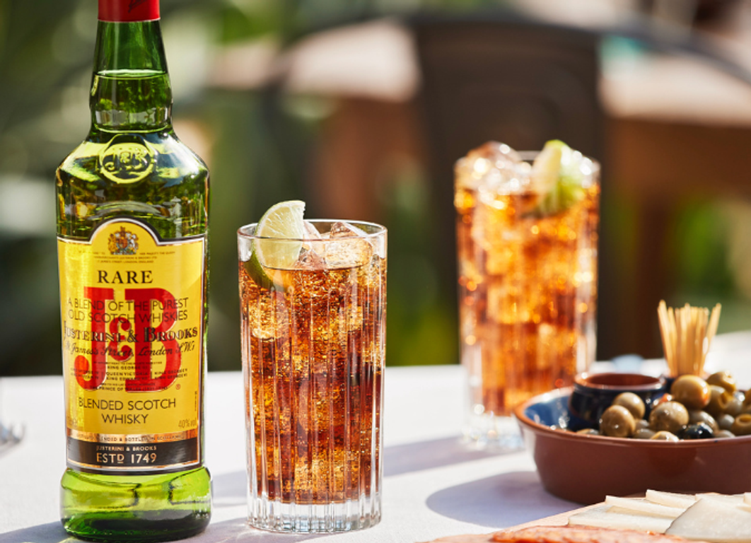 Botella de whisky J&B sobre una mesa de terraza con dos cócteles y un plato de aceitunas