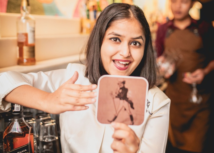 A woman with short, dark hair smiling and holding up a Johnnie Walker placemat whilst leaning on a bottle of Black Label Johnnie Walker whisky behind the bar.