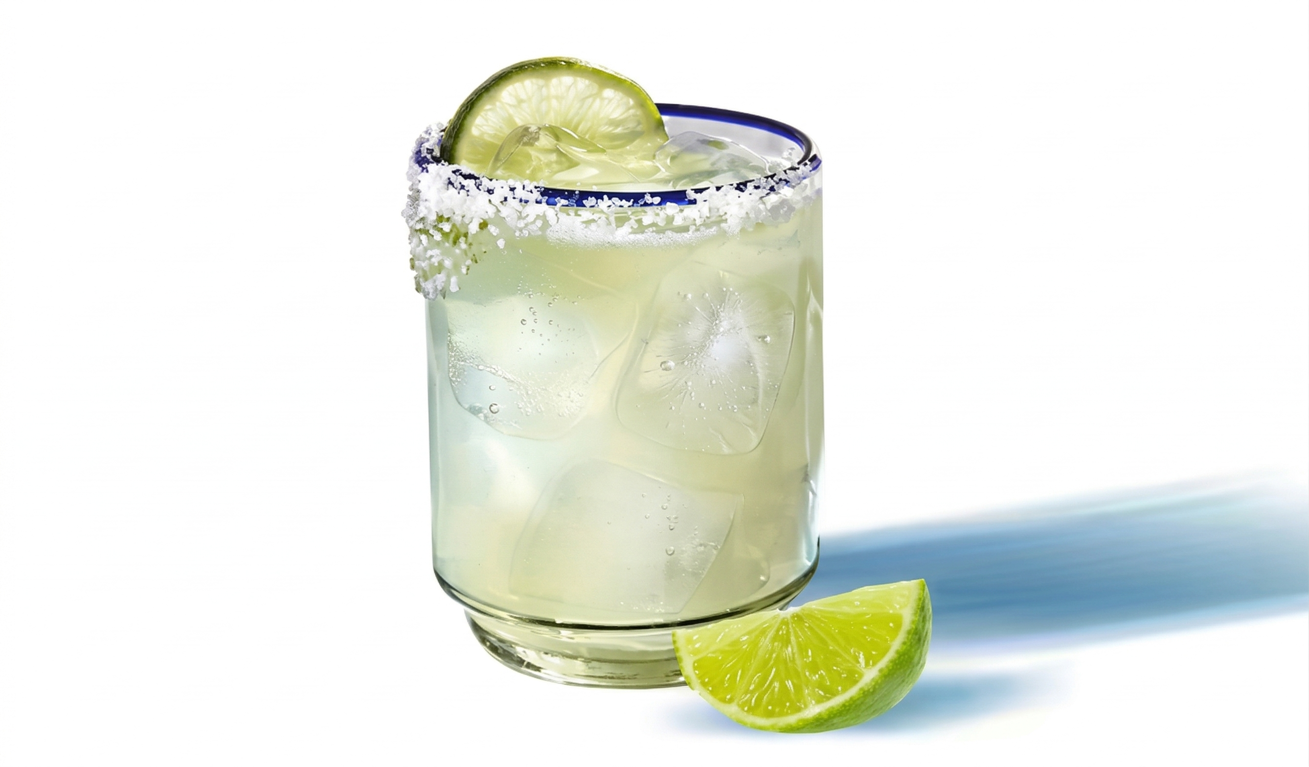 Coquetel tipo Margarita em copo baixo com gelo. Metade da borda possui crosta de sal e é decorada com uma rodela de limão verde. Uma fatia de limão repousa ao lado. Fundo branco isolado com uma longa sombra azul projetada pelo copo.
