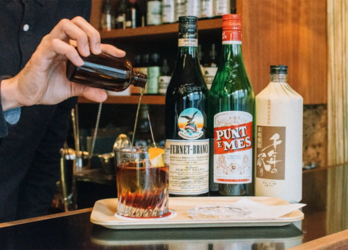 Bartender pouring drink into glass on table beside three bottles of alcohol.   