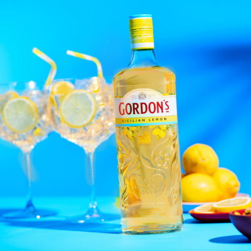 Bottle of Gordon’s Sicilian Lemon gin beside two G&T glasses against blue background 