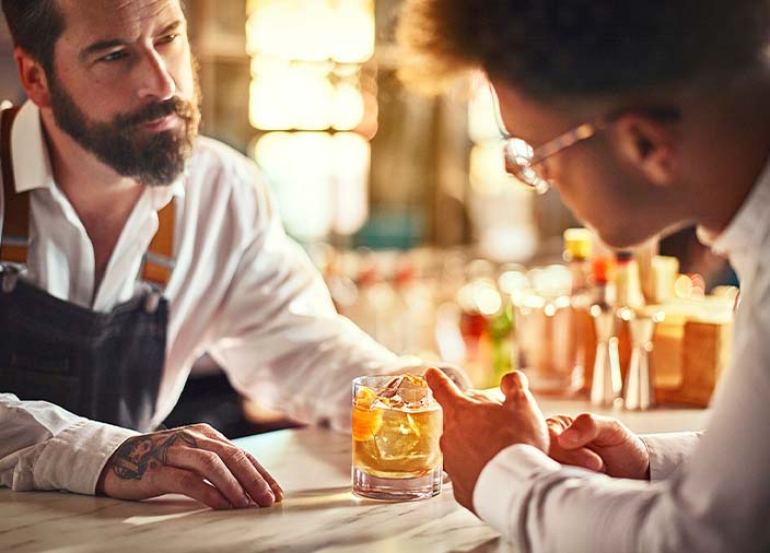 A bar staff member speaking to someone at the bar over a cocktail.  