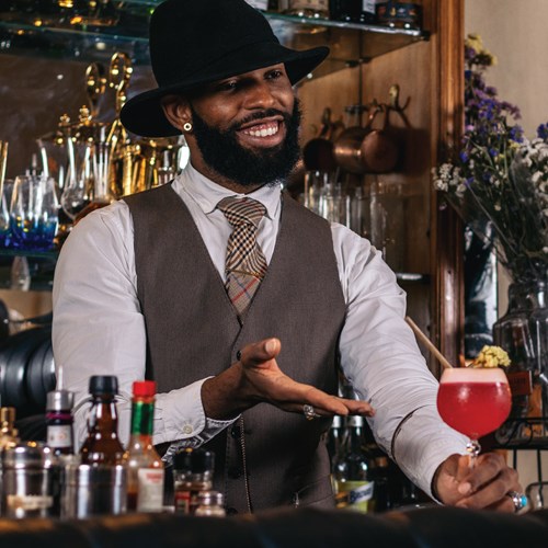 Smiling bartender in a hat presenting a red cocktail with lime garnish.