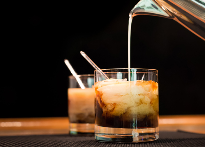 Two glasses of White Russian cocktail, with heavy cream being poured into dark liquid from a small jug.