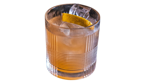 Copo rocks de cristal com drinque âmbar, gelo e twist de casca de limão. Fundo branco.