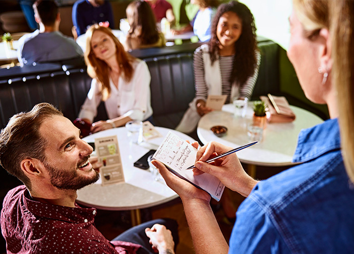  Three people sitting at a table, man in the left corner looks up and smiles at staff member taking down their order on a small notepad, holding a pen. 