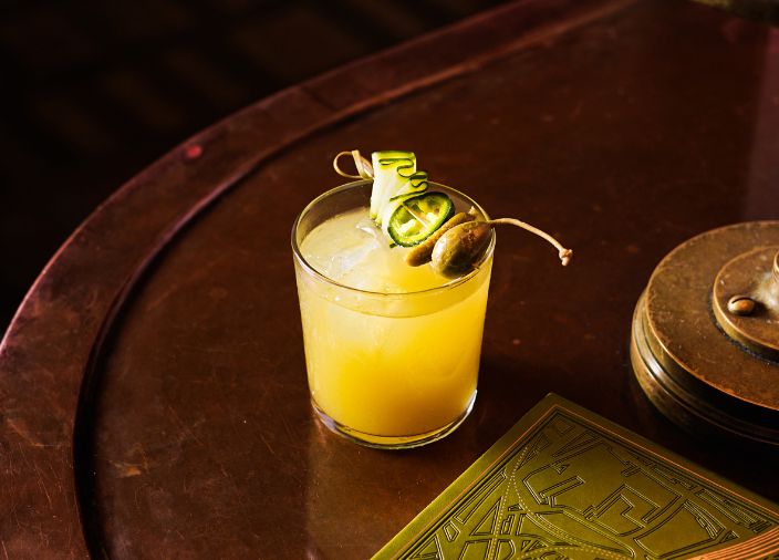 A yellow cocktail with a caperberry and cucumber garnish.