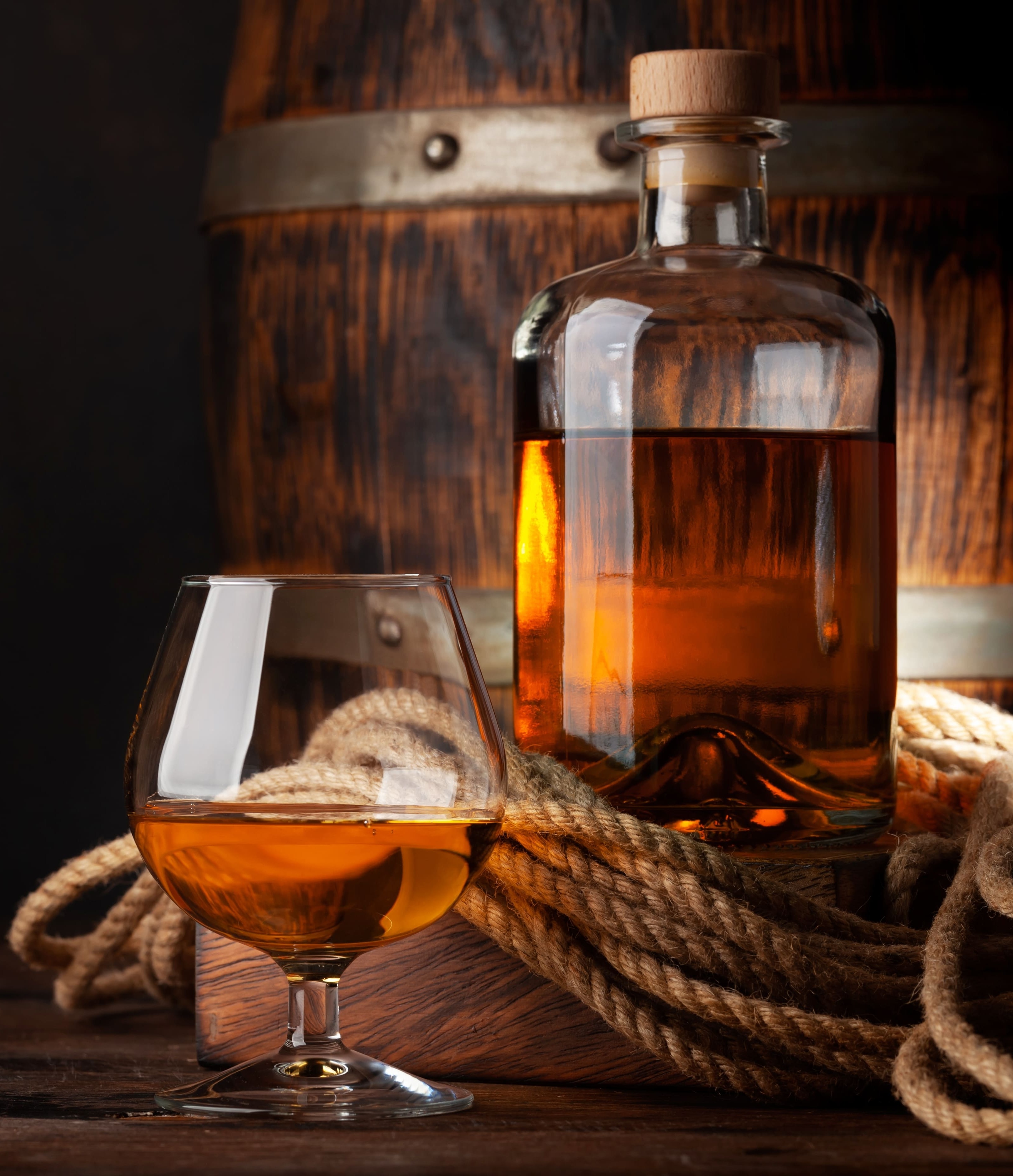 Garrafa de vidro com rolha e taça contendo bebida âmbar, como rum, sobre madeira. Uma corda rústica envolve a base da garrafa. Ao fundo, um barril de madeira escuro. Iluminação dramática destaca o brilho dourado do líquido contra o fundo sombrio.