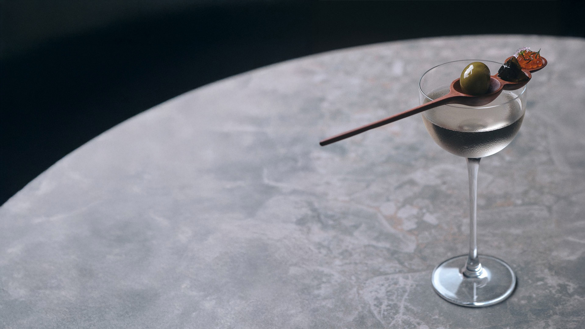 Coquetel transparente em taça suada sobre mesa de mármore cinza. Atravessada na borda, uma colher de madeira decorativa serve uma azeitona verde grande, uma guarnição escura e ovas laranjas. Fundo escuro e elegante.