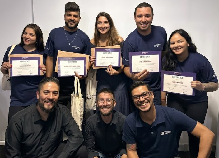 Grupo de oito pessoas sorridentes posando para foto com fundo claro. Na fileira de trás, cinco pessoas em pé seguram certificados. Na frente, três homens estão agachados. A maioria veste camisetas em tom azul-marinho.