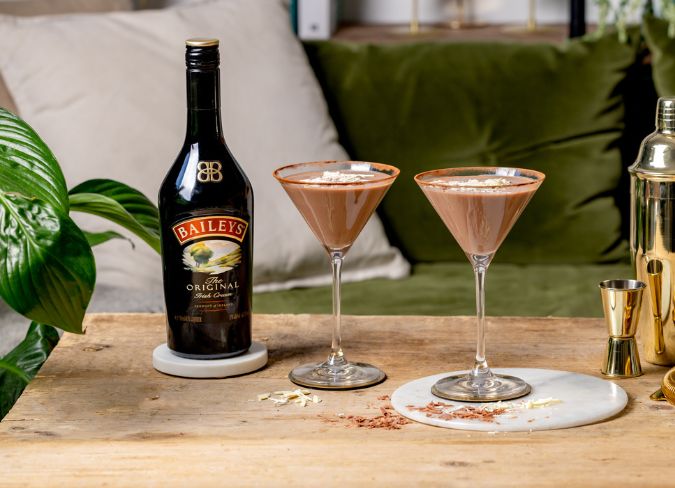 garrafa de baileys ao lado de dois drinks servidos em taças e guarnecidos com grãos de café