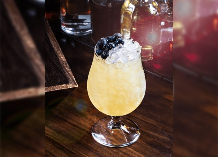 Light yellow cocktail filled with ice and topped with blueberries on a wooden tabletop.