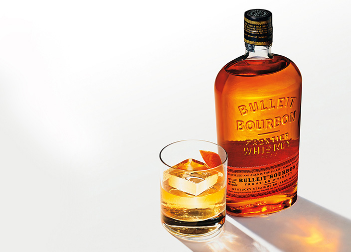  A bottle of Bulleit Bourbon on a white table next to a tumbler glass filled with Bulleit and a large cube of ice. Bright shadows are cast to the bottom right corner.