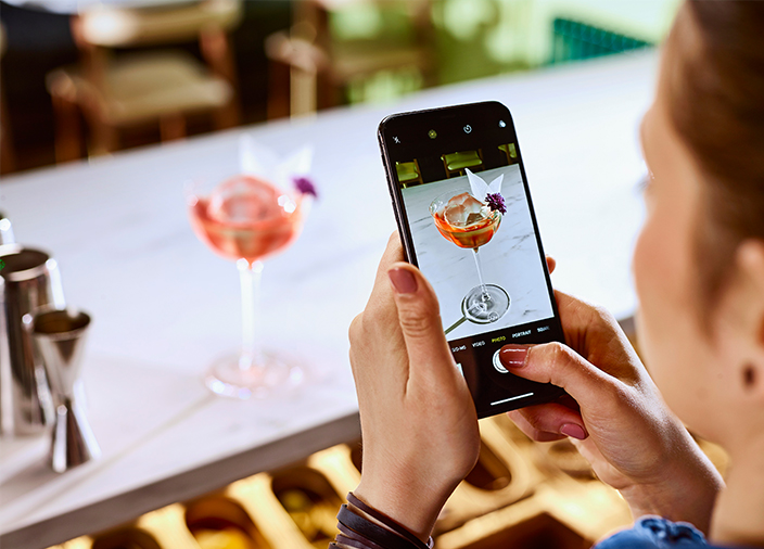 A hand holding a smartphone in camera mode pointed at a cocktail on the bar. The pink cocktail is garnished perfectly with flowers in different colours.